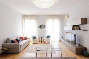 A modern living room with a grey sofa, two white armchairs, a coffee table, a TV, and a bookshelf. - Riviera di Chiaia 207 - 7° Piano (Naples)