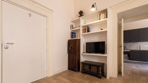 Ein Zimmer mit Holzboden im Fischgrätmuster, einer weißen Tür, einem Holzkabinett, einem Fernseher auf einem Regal und einem durch einen offenen Durchgang einsehbaren Küchenbereich.