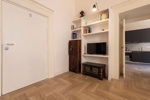 Ein Zimmer mit Holzboden im Fischgrätmuster, einer weißen Tür, einem Holzkabinett, einem Fernseher auf einem Regal und einem durch einen offenen Durchgang einsehbaren Küchenbereich.