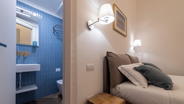 Ein Badezimmer mit Waschbecken, Spiegel und Handtuchhalter. Ein Nachttisch mit Lampe und ein Bett mit Kissen.