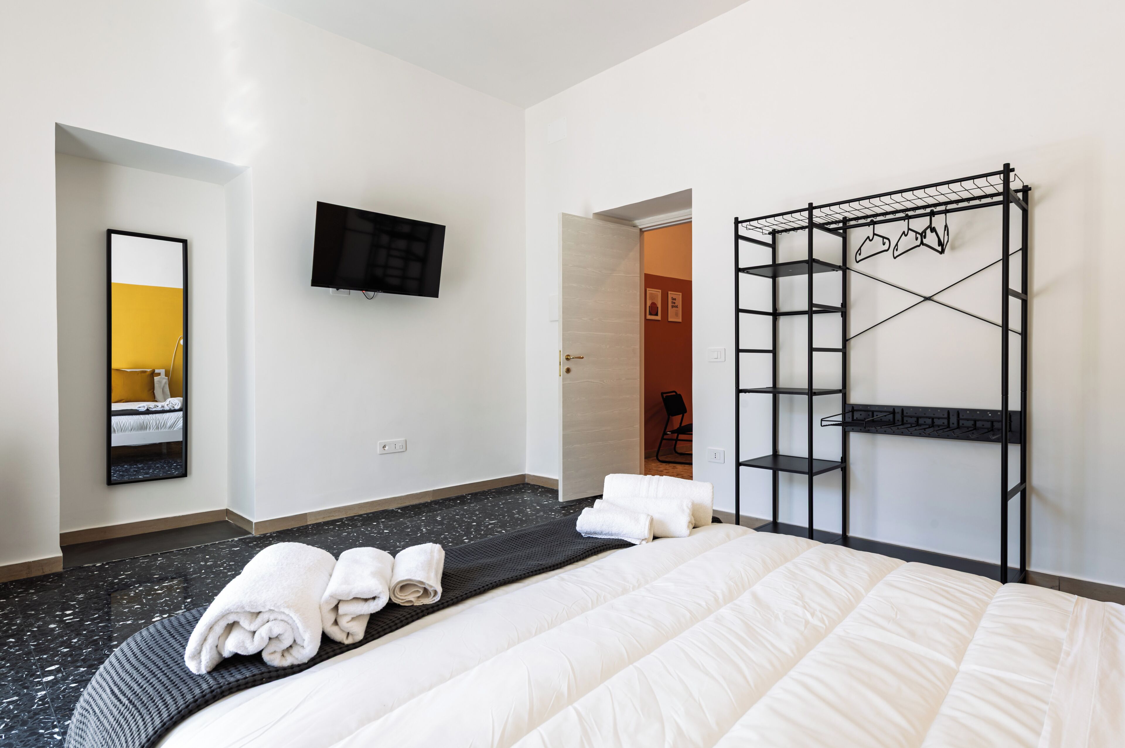 Ein Hotelzimmer mit einem Bett, einem Fernseher, einem Spiegel, einem schwarzen Metallregal und einer Tür zu einem anderen Raum.