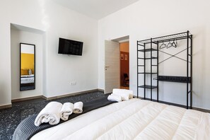 Une chambre d’hôtel comprenant un lit, une télévision, un miroir, une étagère en métal noir et une porte donnant sur une autre pièce.
