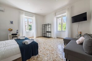 Quarto com cama, sofá, televisão, escrivaninha e janela.