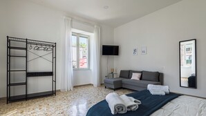 Ett hotellrum med en säng, en grå soffa, en tv, ett metallställ med kläder och ett fönster med gardiner.