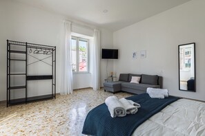 A hotel room with a bed, a gray sofa, a TV, a metal rack with clothes, and a window with curtains. - Casa Luce del Sole (Naples)