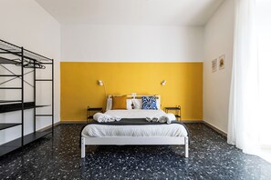 Una habitación con una cama, una pared amarilla, un suelo con estampado blanco y negro y un mueble estantería de metal.