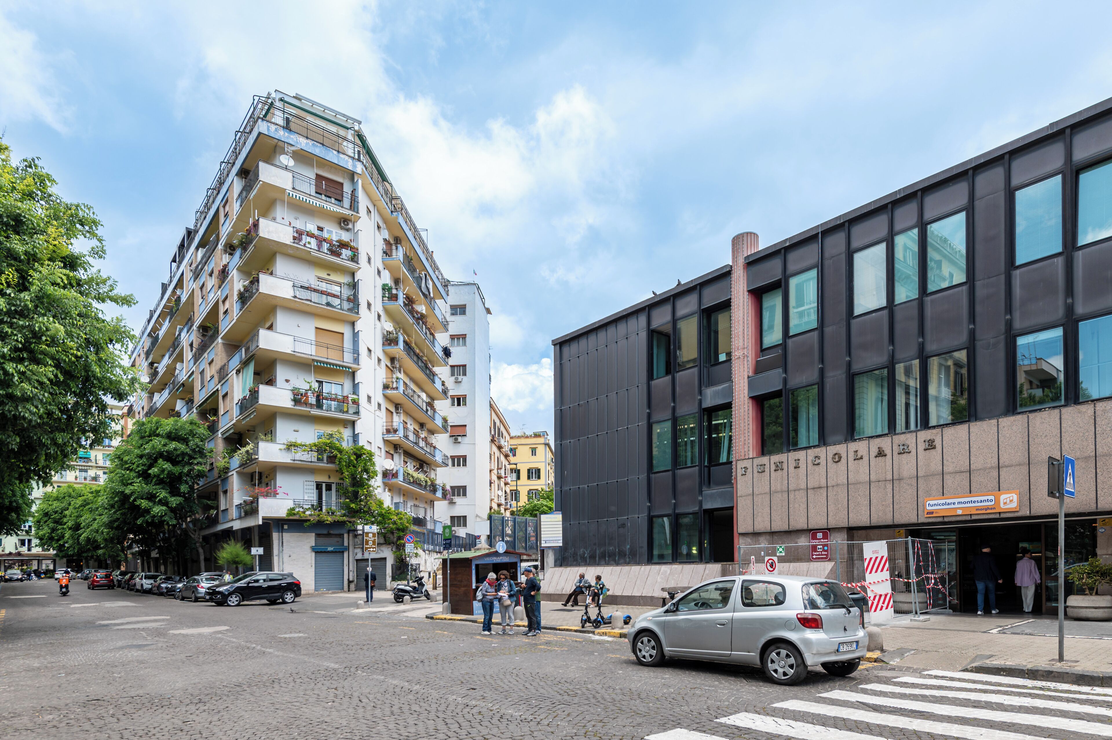 Un edificio a più piani con balconi, un complesso moderno con ampie finestre e un attraversamento pedonale con auto e pedoni.