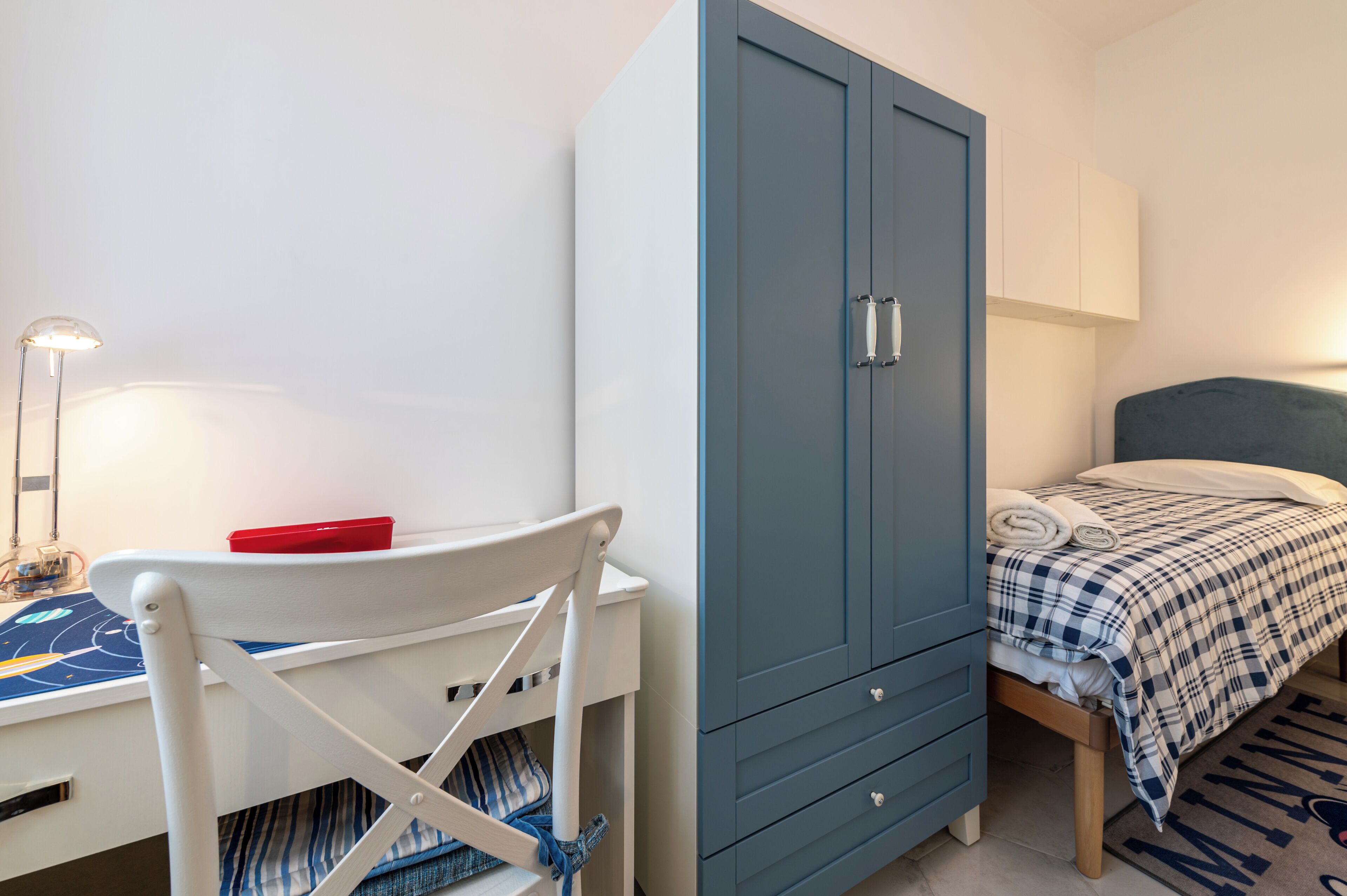 A bedroom with a bed, a blue wardrobe, a desk with a chair, and a lamp.