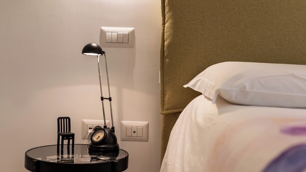 A neatly made bed with white linens, a black bedside lamp, and a small table with a clock and a bottle. - La Favola di San Martino (Naples)