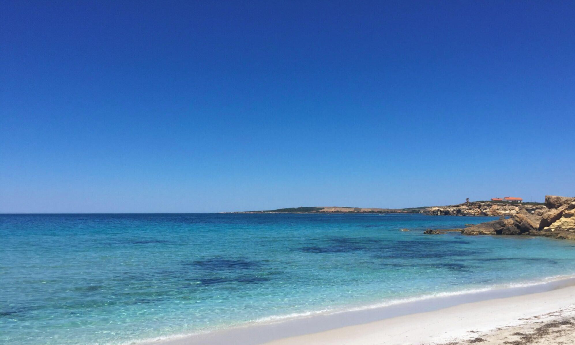 A beach with clear turquoise water, white sand, and rocky outcrops.