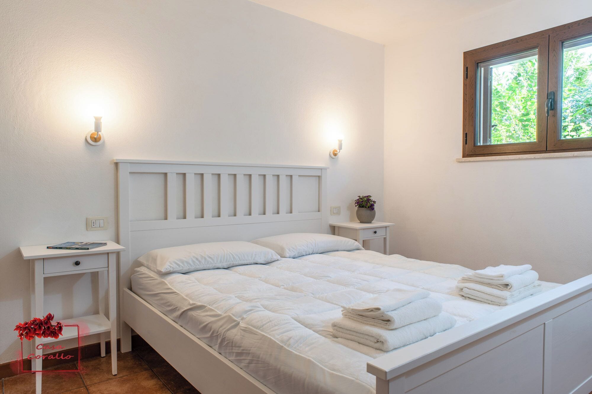 Cama bem arrumada com roupa de cama e almofadas brancas, mesa de cabeceira com abajur, uma pequena planta e uma janela com vista para o verde.