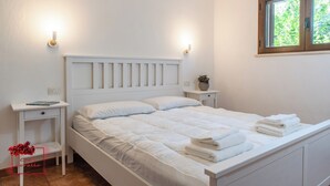 A neatly made bed with white bedding and pillows, a bedside table with a lamp, a small plant, and a window with a view of greenery. - Casa Corallo (Baratili San Pietro)