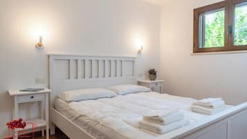 Cama bem arrumada com roupa de cama e almofadas brancas, mesa de cabeceira com abajur, uma pequena planta e uma janela com vista para o verde.