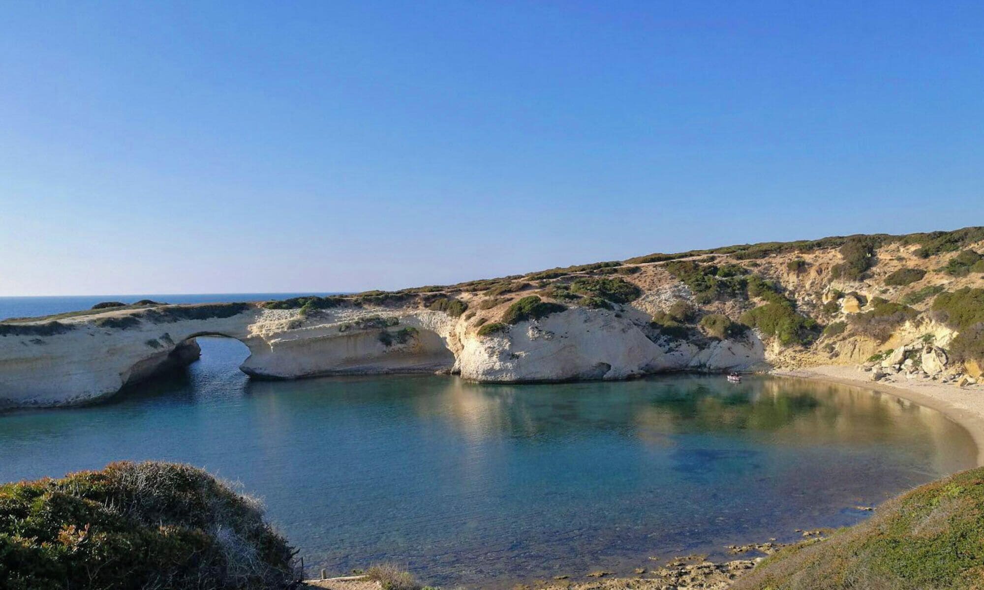 Paisagem costeira com uma formação rochosa em arco natural, uma praia de areia e um céu azul límpido.