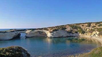 Paisagem costeira com uma formação rochosa em arco natural, uma praia de areia e um céu azul límpido.