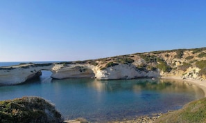Paisagem costeira com uma formação rochosa em arco natural, uma praia de areia e um céu azul límpido.