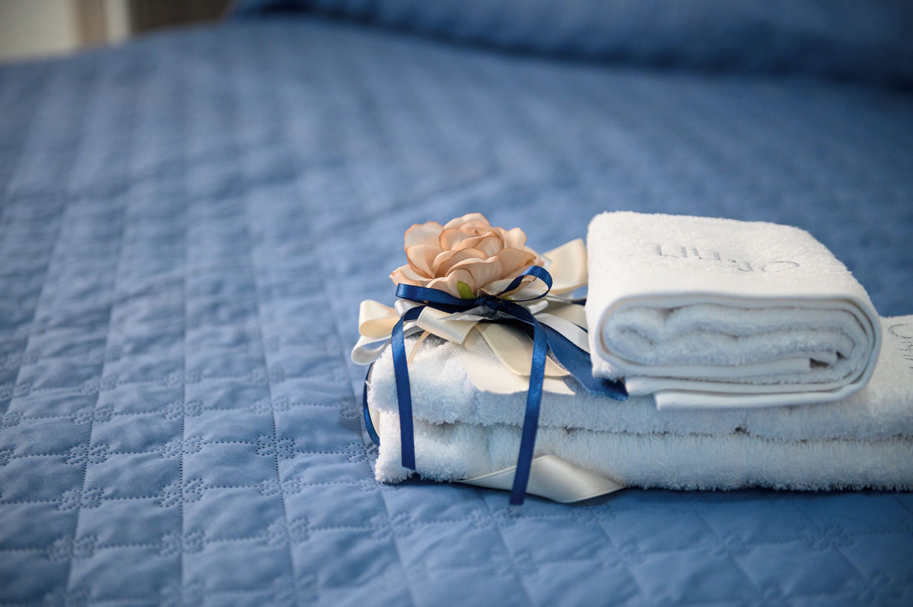 A neatly folded white towel with a blue ribbon and a peach-colored flower on a blue quilted bedspread.