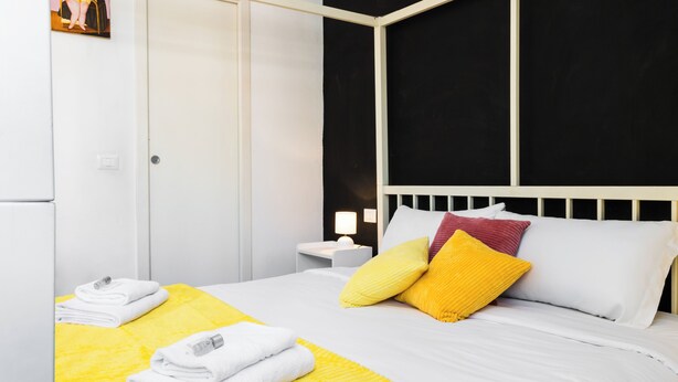 Una cama bien hecha con ropa de cama blanca y amarilla, un cabecero blanco y una mesita de noche pequeña con una lámpara.