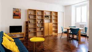 Una sala de estar con un sofá azul, una mesa redonda amarilla, estanterías de madera, un televisor y un comedor con sillas.
