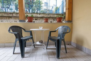 A balcony with two plastic chairs and a small table, potted plants on the ledge, and a view of a blue fence and trees. - Civico 79 (Naples)