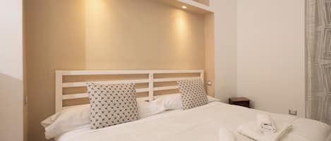 Un lit bien fait, avec du linge de lit blanc et deux coussins à motifs, une petite table de chevet et un luminaire fixé au mur au-dessus du lit.