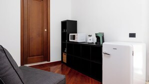 Ein Zimmer mit Holztür, einer schwarzen Regalkonstruktion mit Mikrowelle und Kaffeemaschine, einem weißen Kühlschrank und einem grauen Sofa.
