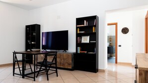 Ein modernes Wohnzimmer mit einem Flachbildfernseher, einem Esstisch mit Stühlen, einem schwarzen Bücherregal und einem Holzschrank.