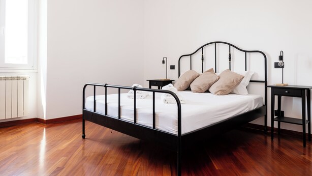 Ein Einzelbett mit schwarzem Metallgestell, weißer Bettwäsche und beigen Kissen. Ein kleiner Nachttisch mit Lampe und Steckdose steht daneben. Der Raum hat Holzboden und eine weiße Wand.