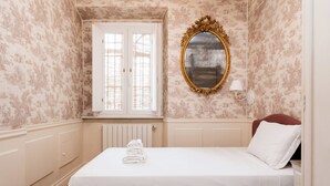 Una cama impecable con sábanas blancas y una toalla doblada, un espejo con marco dorado y una ventana con contraventanas blancas.