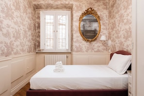 A neatly made bed with white linens and a folded towel, a gold-framed mirror, and a window with white shutters. - Candy Lodge (Rome)