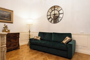 A room with a green sofa, a wooden dresser, a large wall clock, and a lamp. - Candy Lodge (Rome)