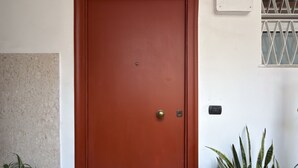 Eine rote, geschlossene Tür mit Schlüsselloch und kleinem Schloss, eine Topfpflanze links und ein Fenster mit Gitterstäben rechts.