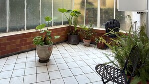 Ein mit Fliesen ausgelegter Balkon mit Topfpflanzen, einem Metallstuhl und einem Fenster mit einem Insektengitter.