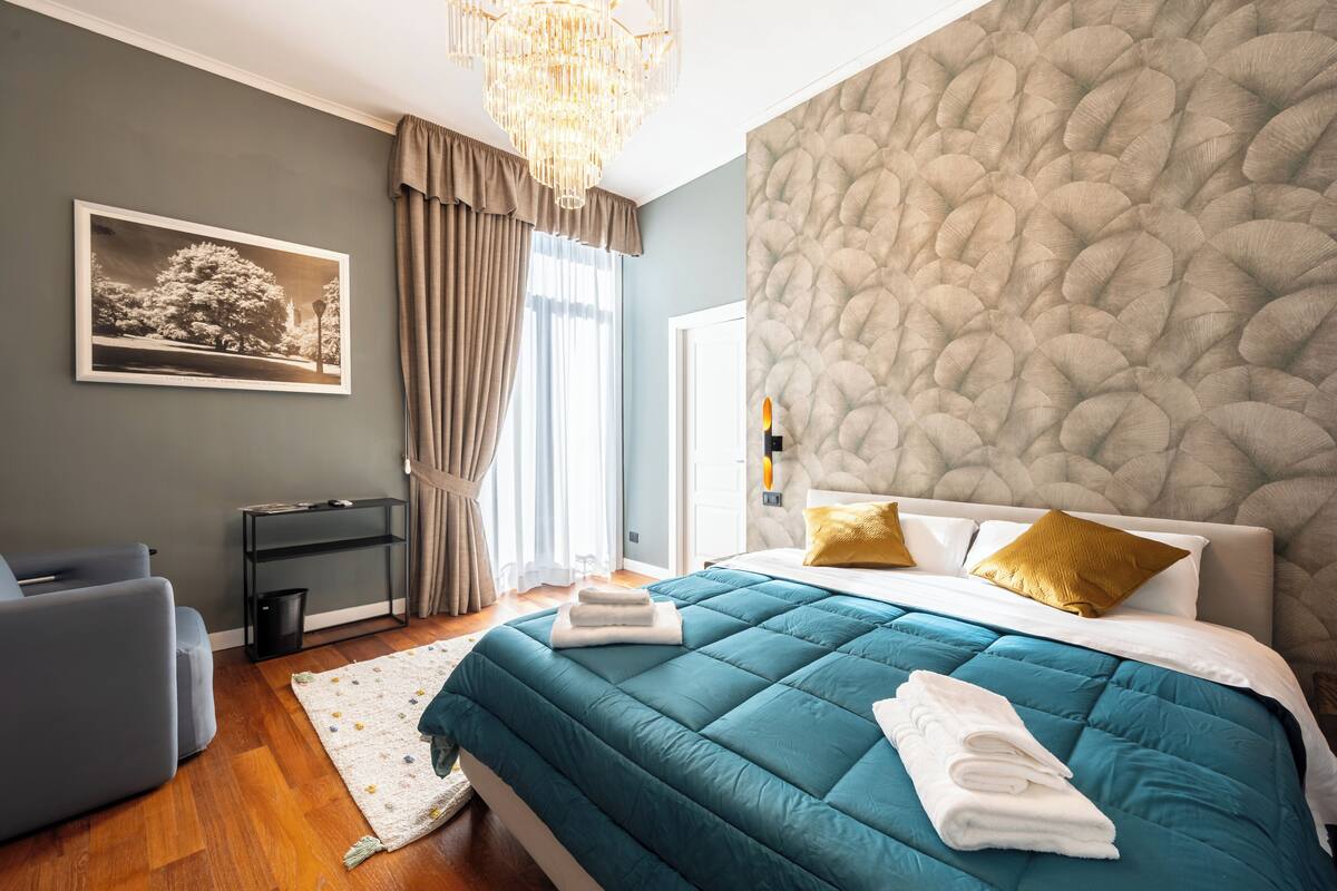 Habitación de hotel con cama, escritorio, silla, candelabro y una obra de arte en la pared.