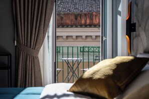 Et hotelværelse med en seng, et vindue med gardiner, udsigt til en balkon med gelænder og et lille bord med stole udenfor.