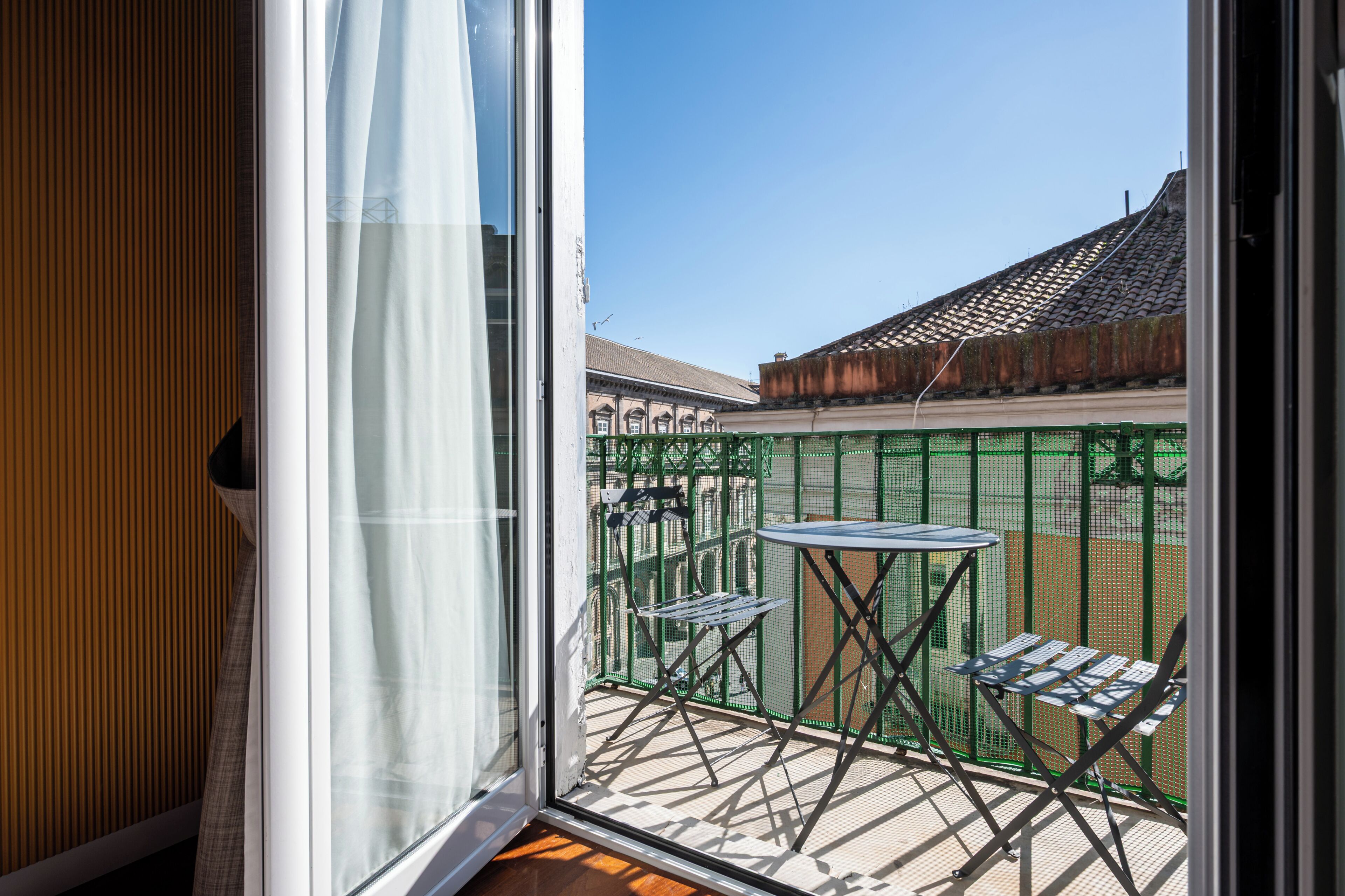 A balcony with a table and chairs, a view of a building with a tiled roof, and a clear sky.