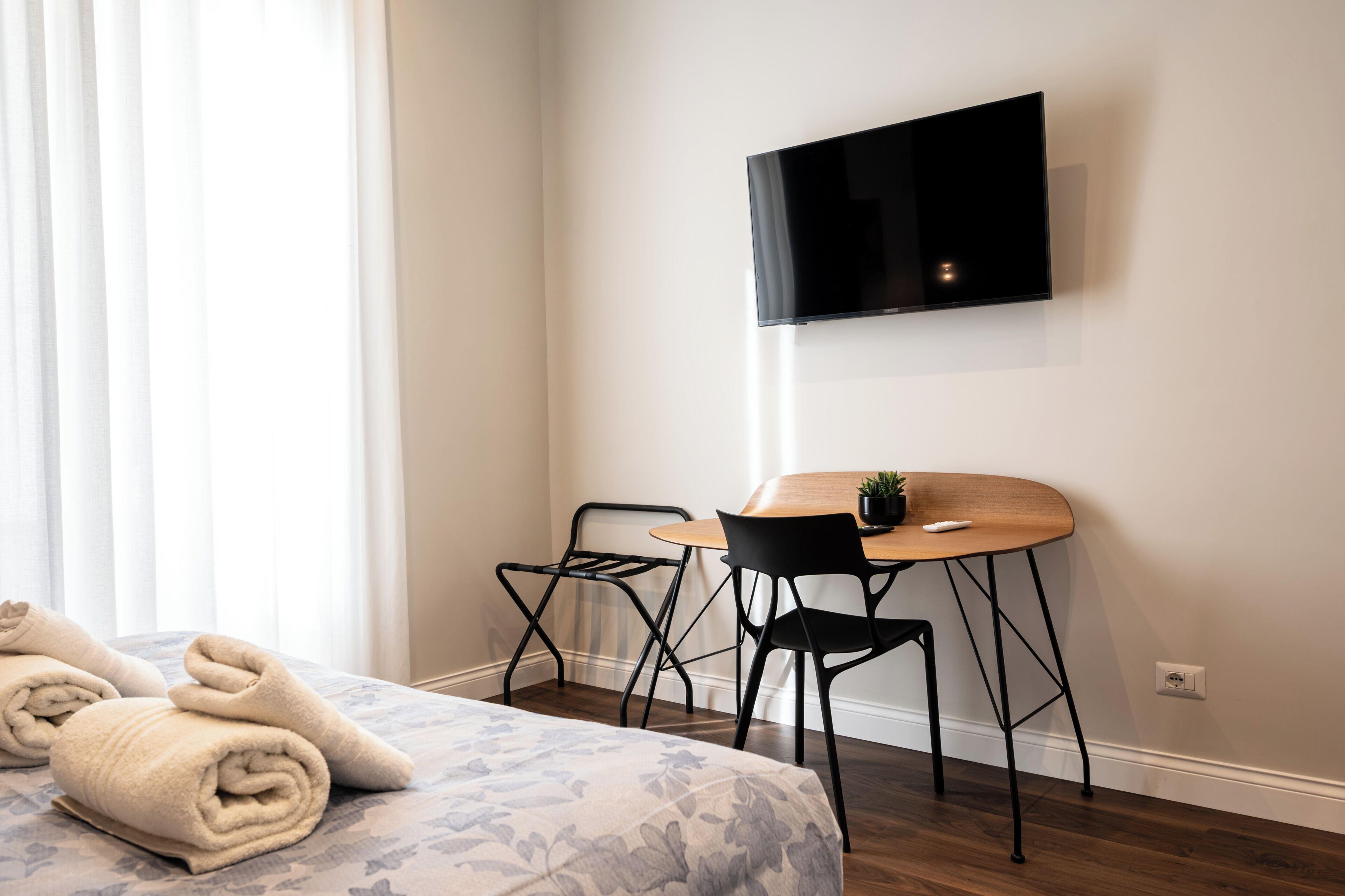 Habitación de hotel con cama, escritorio y silla, televisor montado en la pared y ventana con cortinas.