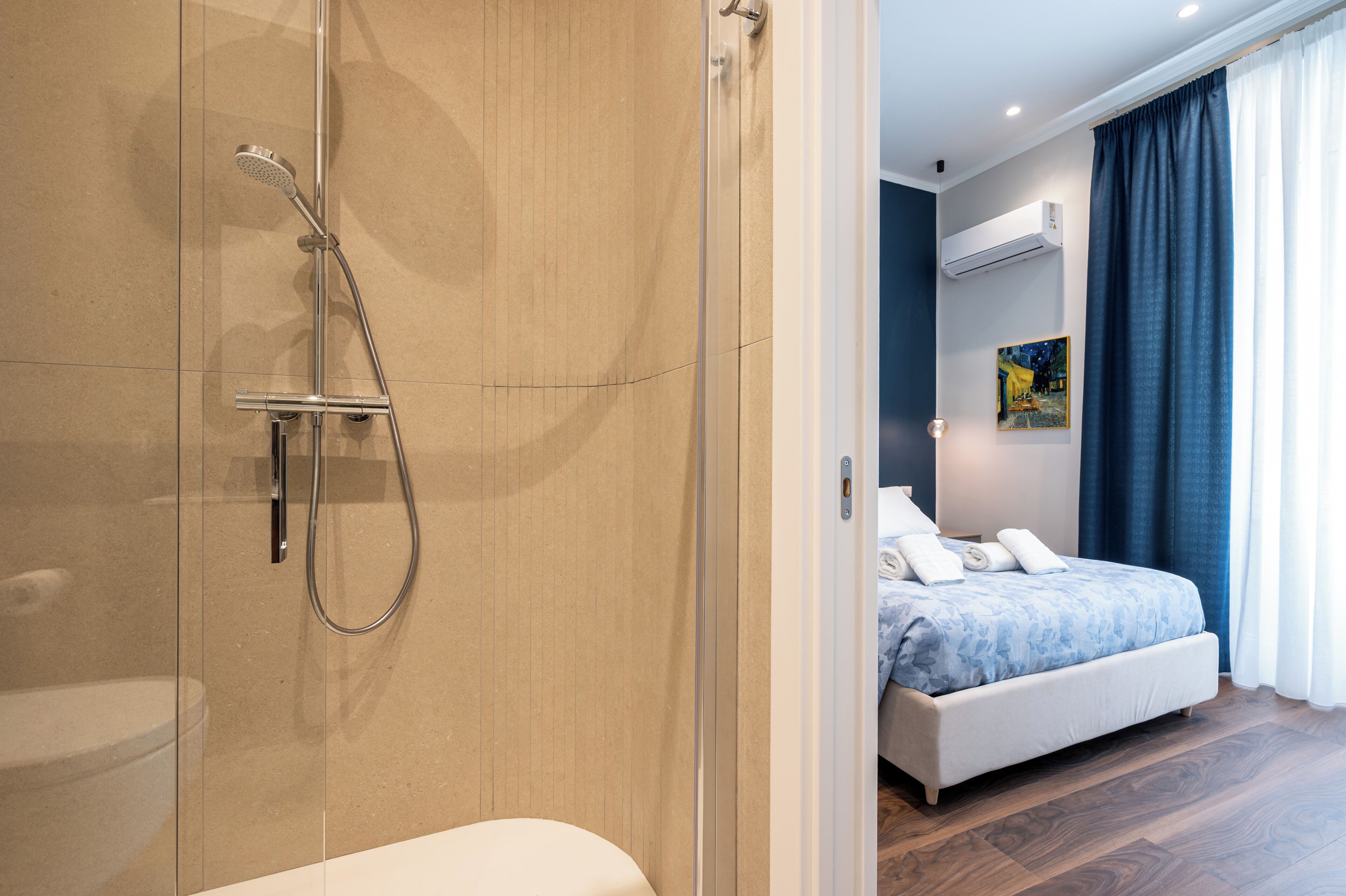 A modern bathroom with a glass shower enclosure, a white toilet, and a bed with a blue bedspread in the adjacent room.