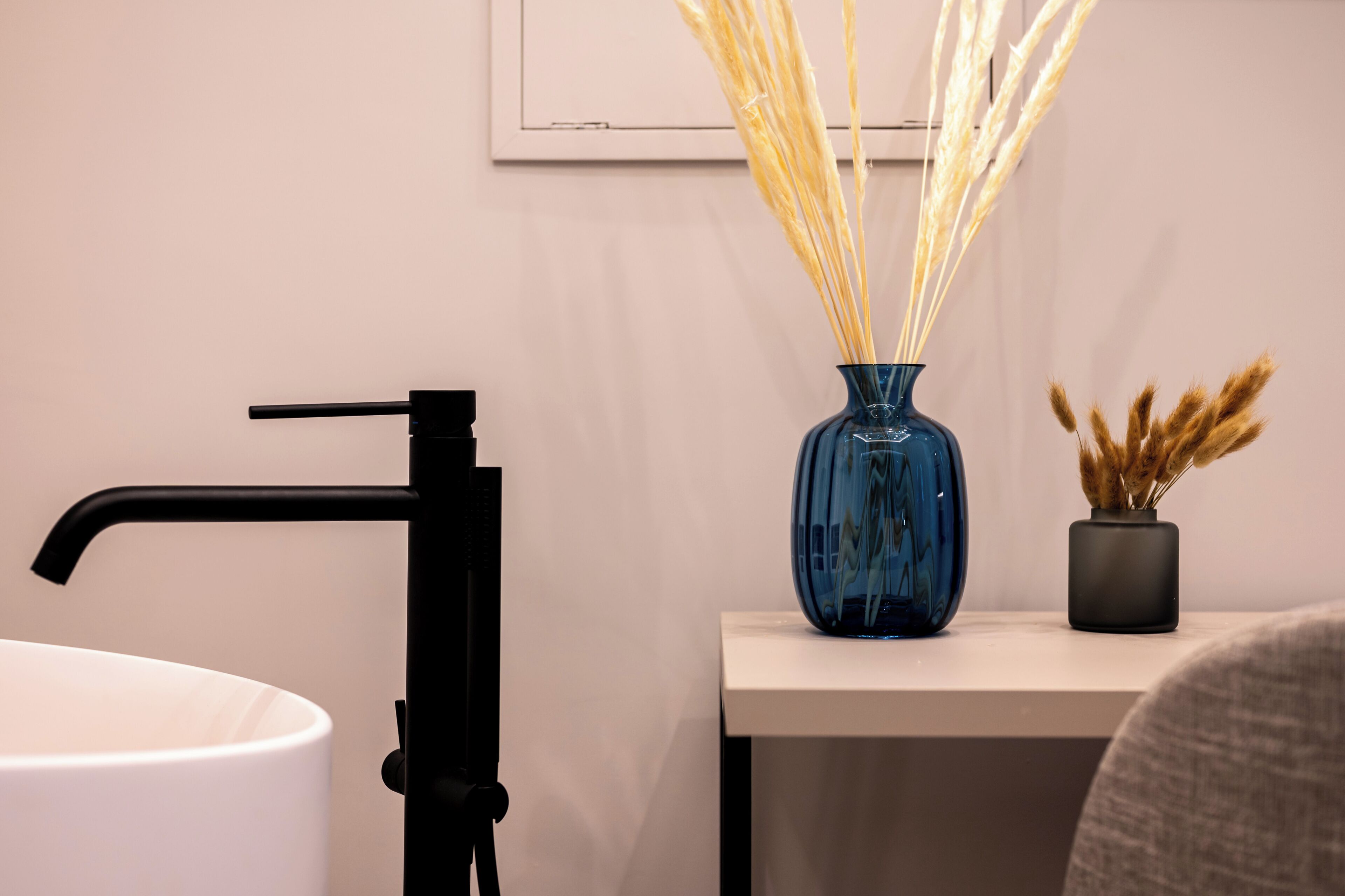 Un baño con un lavabo blanco, un grifo negro y un florero azul con plantas secas sobre una encimera blanca.