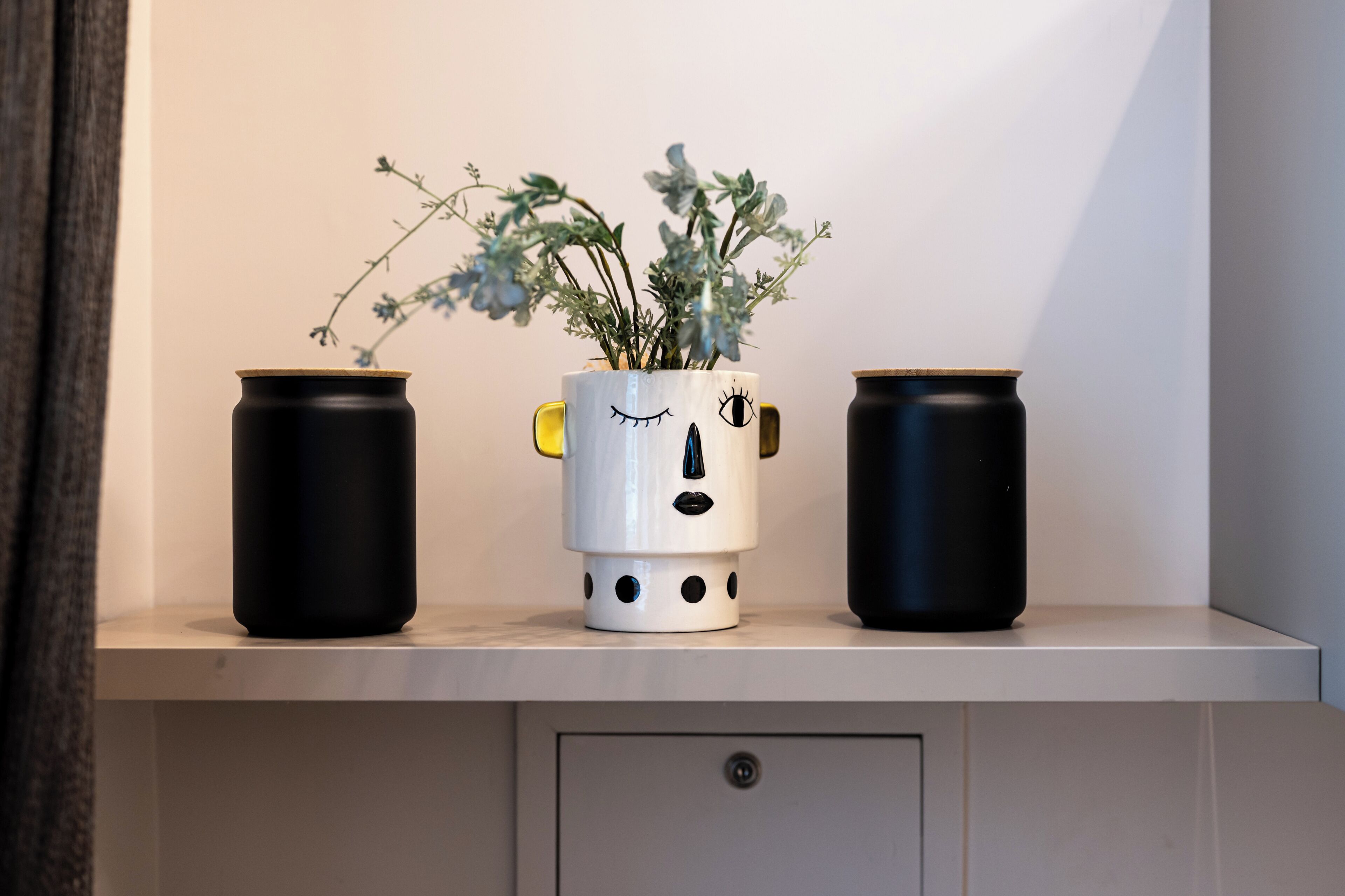 A shelf with three containers, one with a face design and a plant, and two plain black containers.