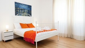 A neatly made bed with white and orange bedding, a white nightstand with a lamp, and a painting of a night scene on the wall. - Dimora Aniene (Rome)