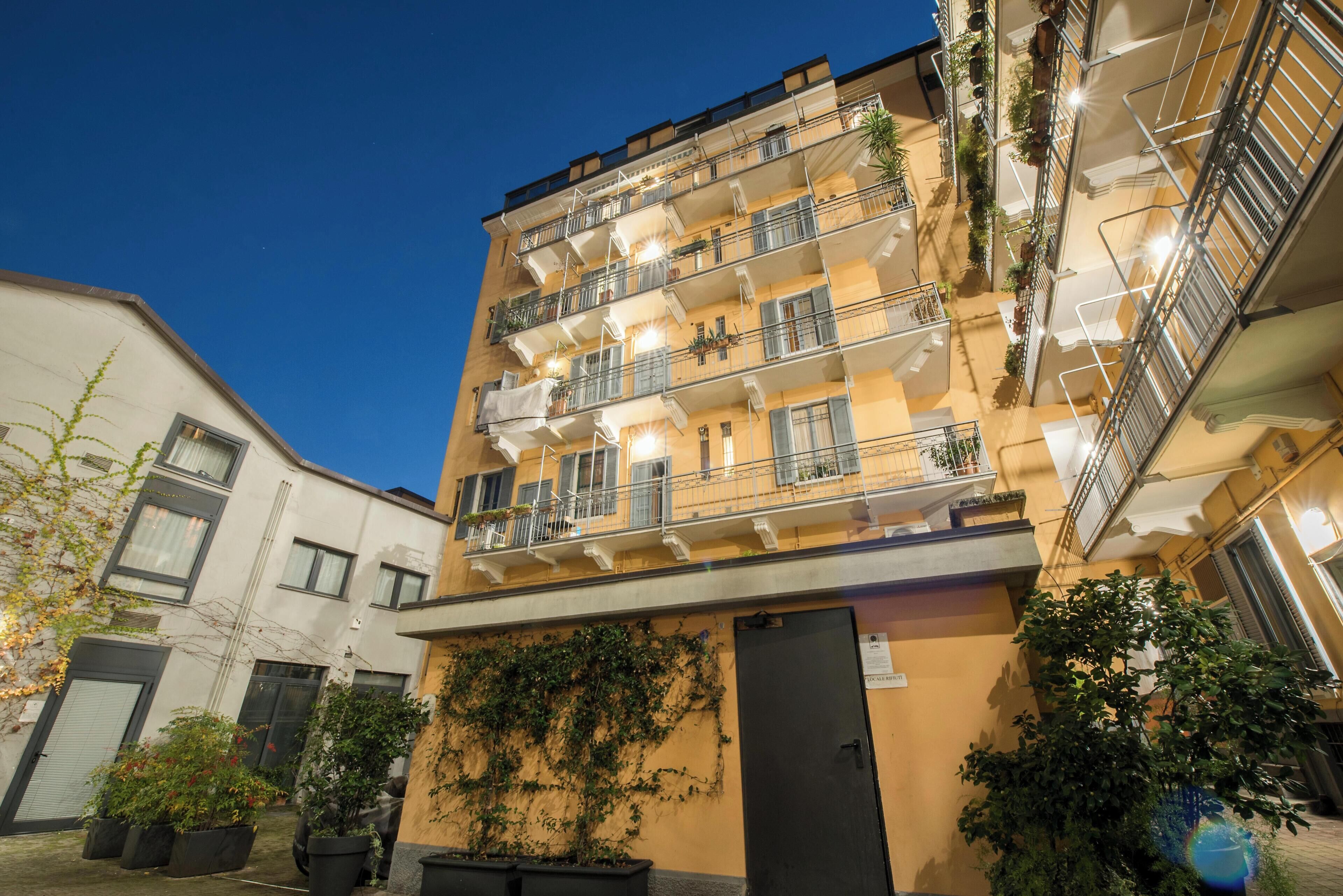 A multi-story residential building with balconies, potted plants, and a door with a sign.