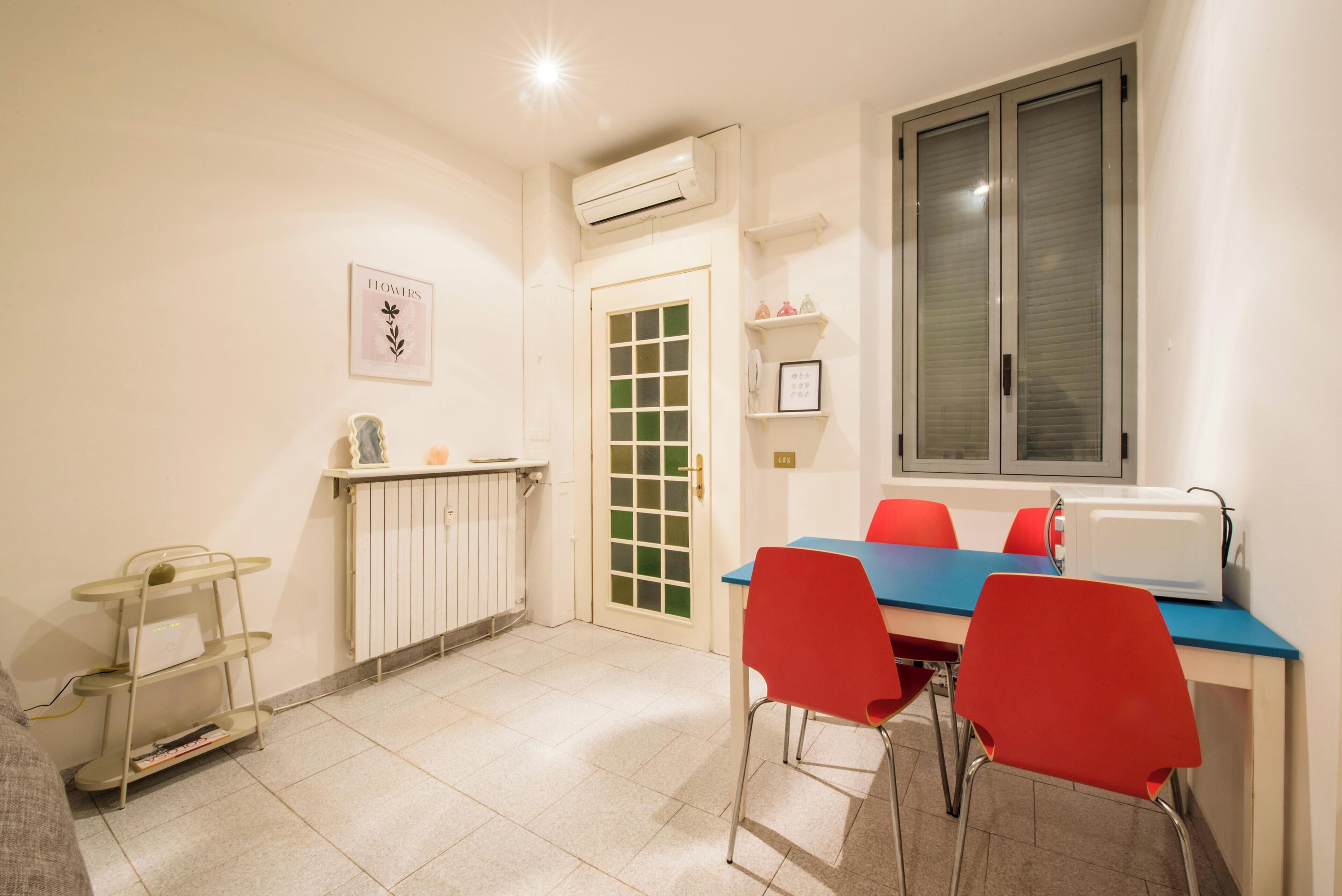 Een kleine keuken met een eettafel, rode stoelen, een witte radiator en een raam met jaloezieën.