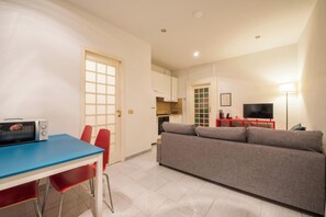 Ein kompakter Wohnbereich mit Küchenzeile, einer Essecke mit blauem Tisch und roten Stühlen, einem grauen Sofa und einem Fernseher auf einem roten Schrank.