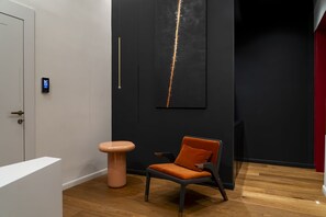A modern interior with a wooden floor, a white door, a digital lock, a tall black wall with a crack, and a chair with an orange cushion. - Via Chiaia 123 - Camera Oltremare (Naples)