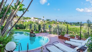 Piscina com espreguiçadeiras, rodeada por vegetação e vista para o mar.