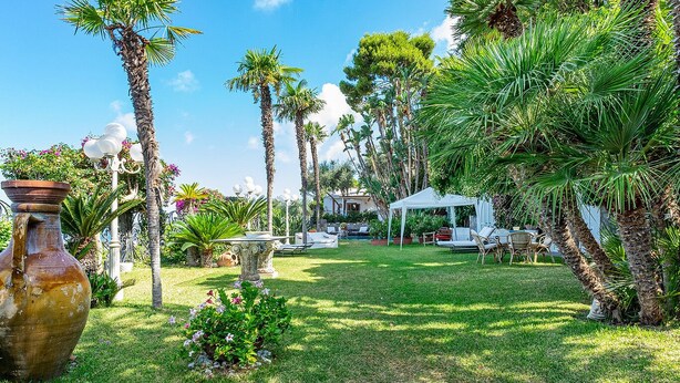 Um jardim bem cuidado, com palmeiras, um gazebo branco e uma mesa de pedra.