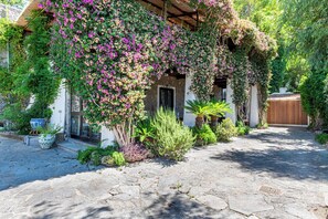 Un patio empedrado con una casa cubierta de enredaderas floreadas, un portón de madera y diversas plantas.