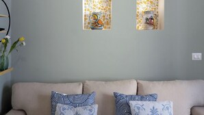 Ein beiges Sofa mit zwei Zierkissen, eine Wand mit zwei Mosaik-Leuchten und eine kleine Vase mit Blumen auf einem Regal.