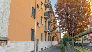 Una stretta via cittadina con edifici a più piani, una recinzione in metallo e un albero con foglie autunnali.